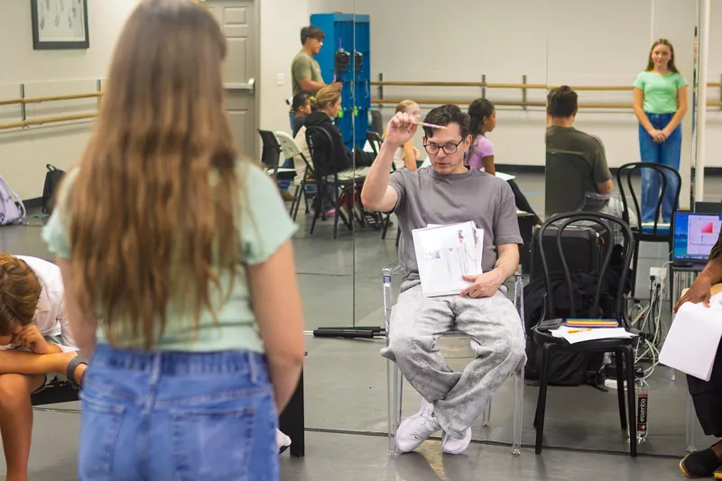 Paul Becker seated on a clear chair in a mirrored studio, pen raised, giving a note to a student standing in front of him while other students watch from chairs
